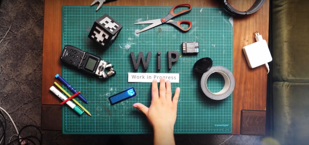 Draufsicht auf Tischarbeitsplatz mit Werkzeugen und eine Hand, die den Schriftzug "WIP - Work in Progress" auflegt