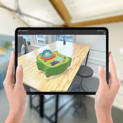 Hände halten Tablet, auf dem ein Tisch abgebildet ist. Auf dem Tablet ist digitales Objekt eingeblendet, das sich scheinbar auf dem Tisch befindet.