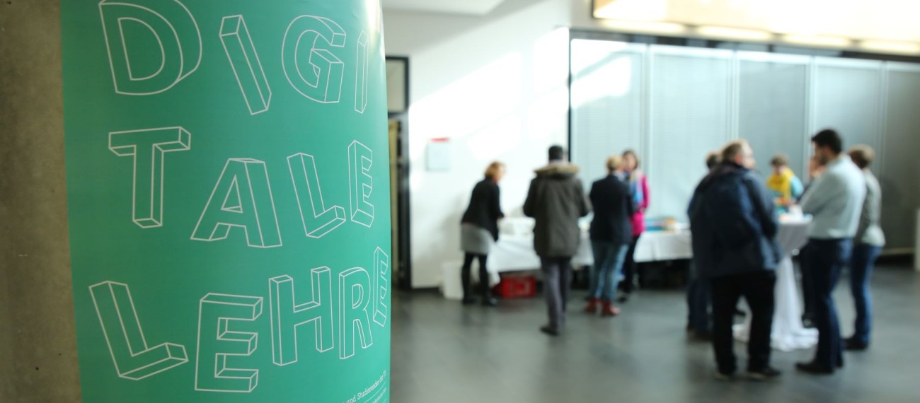 Foto: Ein türkisfarbenes Plakat mit der Aufschrift "Digitale Lehre" ist im Vordergrund zu sehen, während im Hintergrund eine Gruppe von Menschen bei einem Workshop zusammensteht.