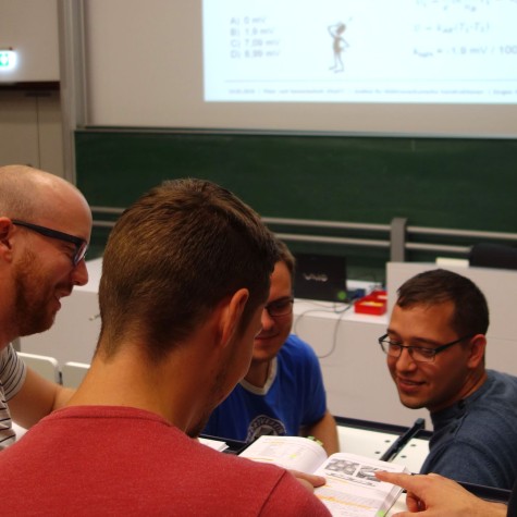 Foto: Eine Gruppe von vier Männern sitzt in einem Klassenzimmer und diskutiert über ein Buch, während im Hintergrund eine grüne Tafel und eine projizierte Präsentation zu sehen sind.