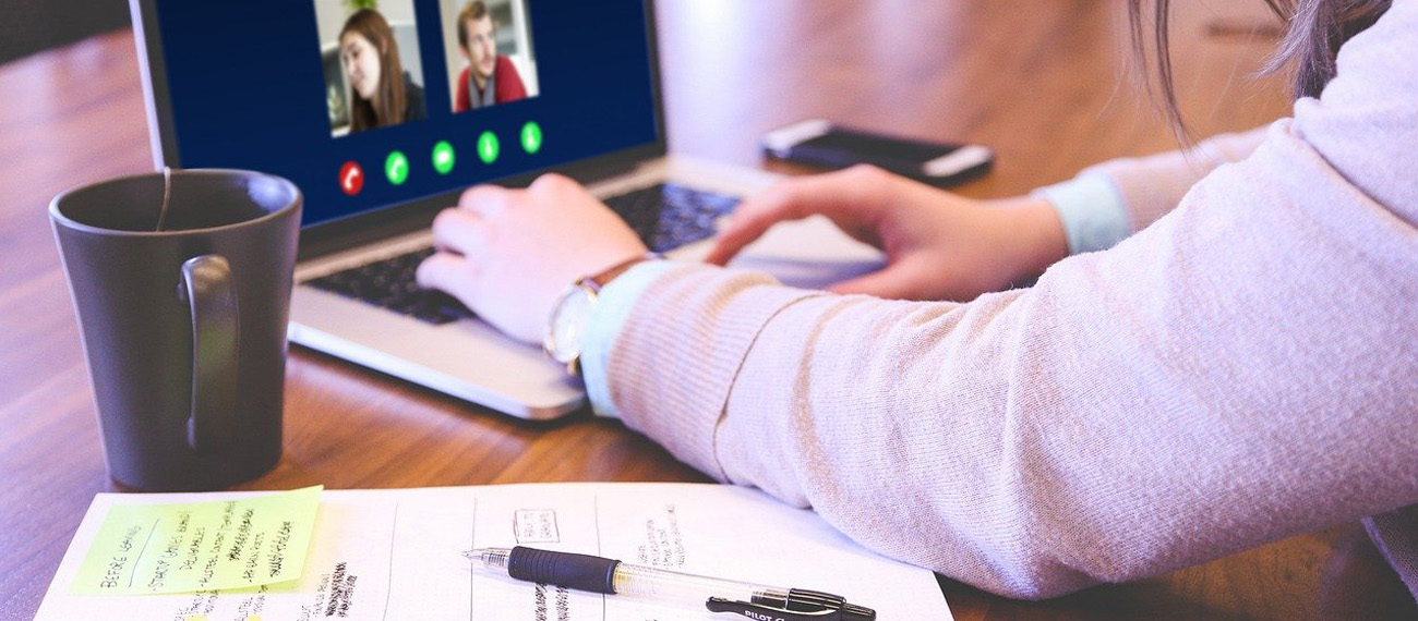 Foto: Eine Person sitzt an einem Schreibtisch und nimmt an einer Videokonferenz auf einem Laptop teil. Auf dem Tisch liegen ein Notizbuch mit Notizen, ein Stift, ein Smartphone und eine Tasse
