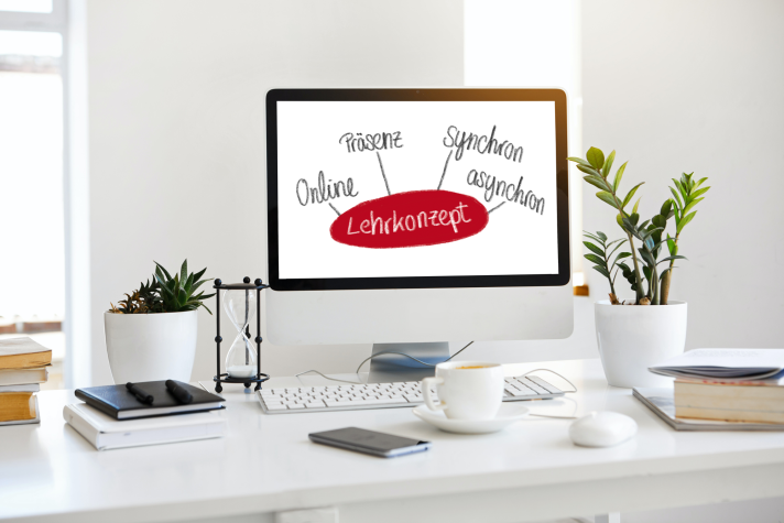 Foto: Ein Arbeitsplatz mit einem Computerbildschirm, auf dem eine Grafik mit dem Wort "Lehrkonzept" in der Mitte und den Begriffen "Präsenz", "Synchron", "Asynchron" und "Online" drumherum angezeigt wird. Der Schreibtisch ist mit Pflanzen, Büchern, einer Sanduhr und einer Tasse Kaffee dekoriert.