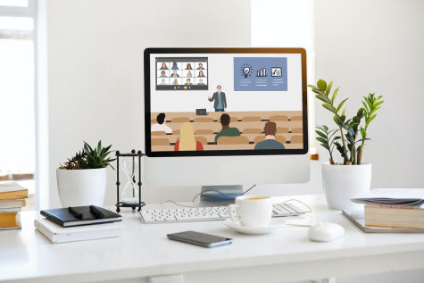 Foto: Schreibtisch mit Monitor, auf dem Hörsaal zu sehen ist, wo Lehrperson eine Präsentation zeigt und weitere Studierende online zugeschaltet sind.