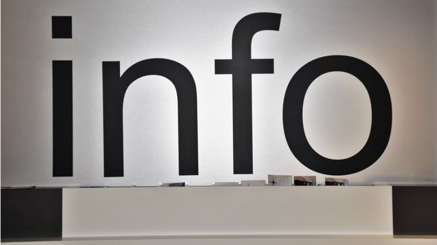Foto: Eine weiße Wand mit dem großen schwarzen Schriftzug "info". Darunter befindet sich eine Theke mit Informationsmaterialien.